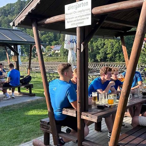 Eine Gruppe junger Männer in blauen T-Shirts sitzt entspannt an einem Tisch im Biergarten und genießt Getränke.