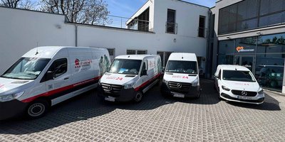 Flotte von Einsatzfahrzeugen vor dem Dienstgebäude Vier Mercedes-Vans stehen vor einem modernen Gebäude, das mit großen Fenstern und einem Willkommensschild ausgestattet ist.