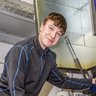 Junger Techniker arbeitet an moderner Maschinenanlage Ein junger Mann in einem grauen Arbeitsanzug lächelt, während er an einer Maschine in einer Werkstatt arbeitet.