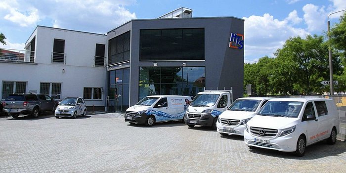 Ein modernes Gebäude mit großen Fenstern und einem klaren blauen Himmel, umgeben von mehreren Lieferfahrzeugen auf einem gepflasterten Parkplatz.