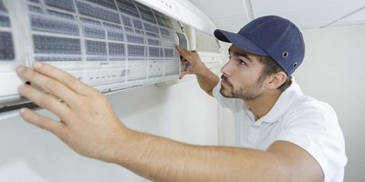 Ein Mechatroniker installiert eine Klimaanlage an der Wand und trägt eine Schirmmütze auf dem Kopf.