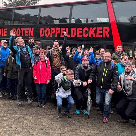 Eine Gruppe von Menschen steht fröhlich vor einem roten Doppeldeckerbus, umgeben von einer bewaldeten Landschaft.