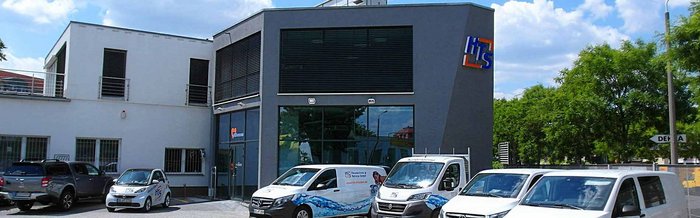 Ein modernes Firmengebäude mit großen Fenstern und einem auffälligen Logo, umgeben von mehreren Fahrzeugen auf einem Parkplatz.