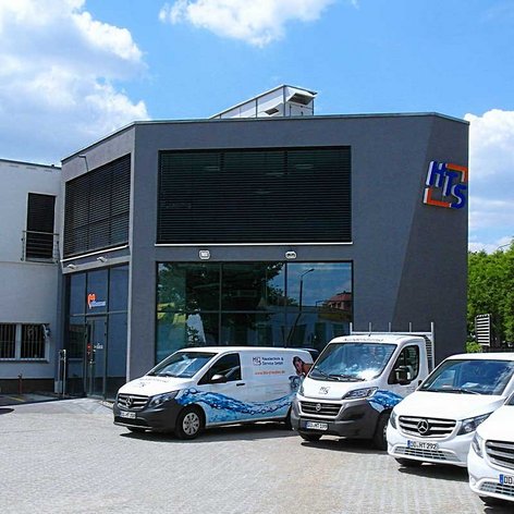 Ein modernes Gebäude mit großen Fenstern und einem klaren blauen Himmel, umgeben von mehreren Fahrzeugen auf einem Parkplatz.