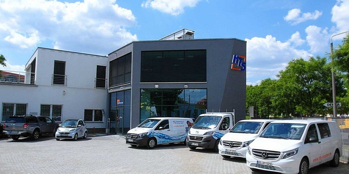 Ein modernes Gebäude mit großen Fenstern und einem klaren blauen Himmel, umgeben von mehreren Fahrzeugen auf einem Parkplatz.