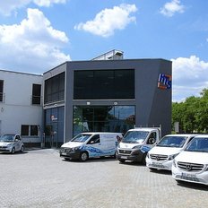Ein modernes Firmengebäude mit großen Fenstern und einem auffälligen Logo, umgeben von mehreren Fahrzeugen auf einem gepflasterten Parkplatz.