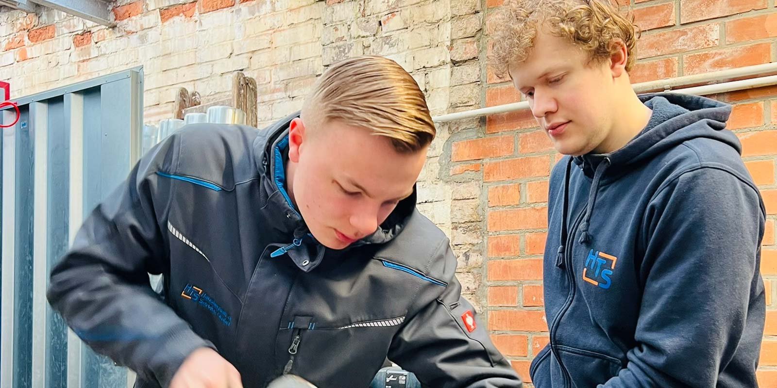 Zwei junge Männer arbeiten in einer Werkstatt, einer konzentriert auf eine Aufgabe, während der andere aufmerksam zuschaut.