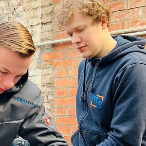 Praktische Ausbildung: Handwerkliche Fähigkeiten im Einsatz Zwei junge Männer arbeiten in einer Werkstatt, einer konzentriert auf eine Aufgabe, während der andere aufmerksam zuschaut.